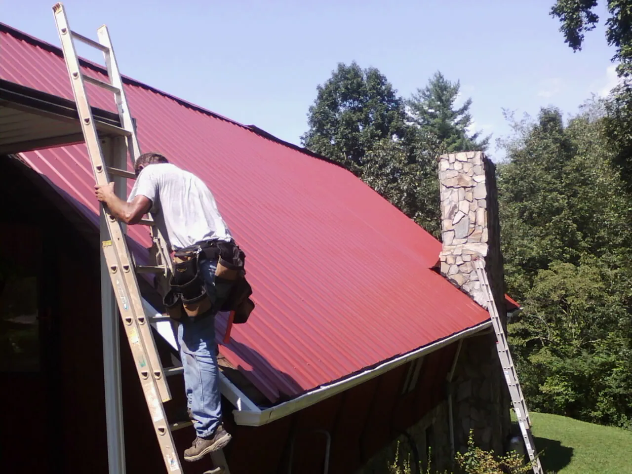 Roofer on a ladder beside a red metal roof and stone chimney — Soffit & Fascia Repair by Roofing Brothers in Eastpointe